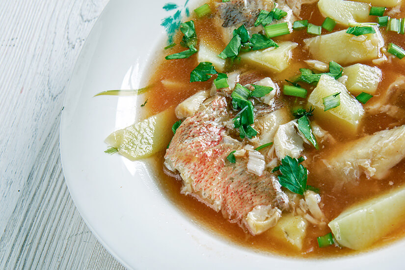Zu erkennen ist eine kräftige Fischsuppe mit Fischstücken, Kartoffeln und frischen Kräutern in einer würzigen Brühe. Gehackte Petersilie und Frühlingszwiebeln dienen als Garnitur. Maltesisch Essen Aljotta