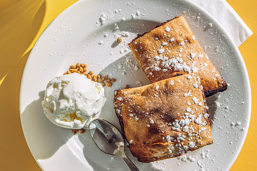 Zu erkennen sind zwei knusprig gebackene Teigtaschen, die mit Puderzucker bestäubt sind. Dazu werden eine Kugel Eis und einige gehackte Nüsse serviert. Malta Essen Imqaret