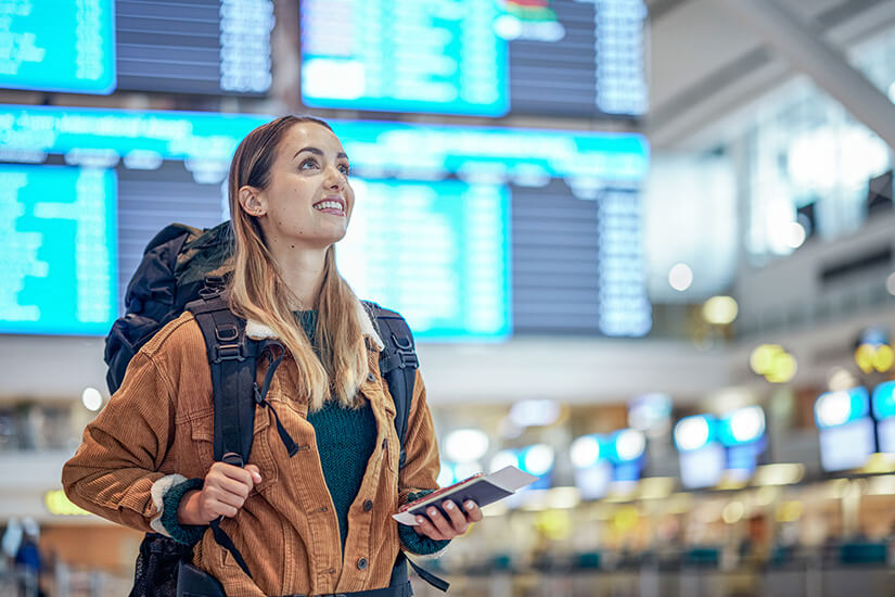 Zu erkennen ist eine Reisende mit Rucksack, Reisepass und Smartphone in einem Flughafenterminal. Im Hintergrund befinden sich große digitale Anzeigetafeln mit Fluginformationen. Low Budget Urlaub Flughafen