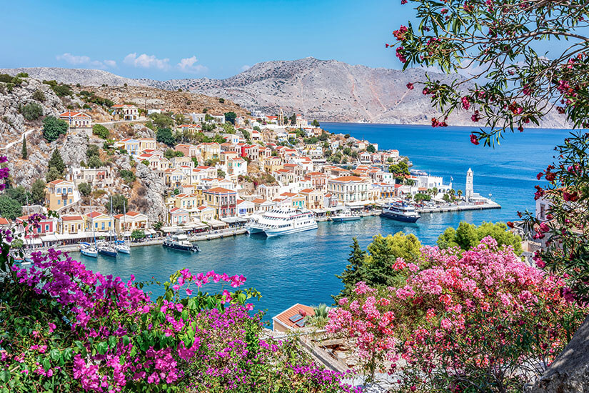 Zu erkennen ist eine malerische griechische Küstenstadt mit pastellfarbenen Häusern, einem Hafen und mehreren Booten. Blühende Bougainvilleen rahmen den Blick auf das tiefblaue Meer und die karge Berglandschaft ein. Spartipps Urlaub Griechenland