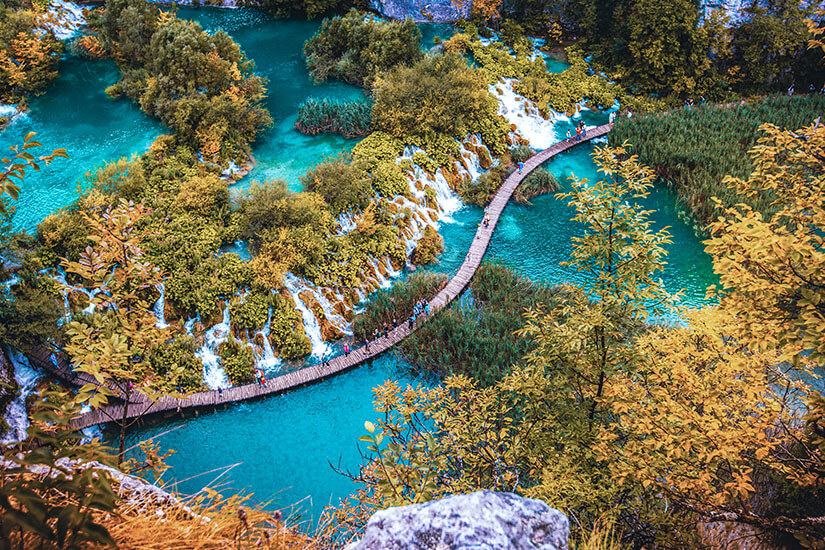 Zu erkennen sind türkisfarbene Seen, zahlreiche kleine Wasserfälle und üppige Vegetation. Ein geschwungener Holzsteg führt durch die eindrucksvolle Wasserlandschaft und wird von mehreren Menschen genutzt. Low Budget Urlaub Kroatien