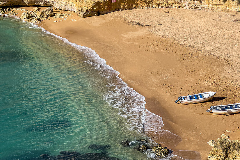 Zu erkennen ist eine kleine, von Felsen eingerahmte Sandbucht mit klarem türkisfarbenem Meer. Zwei kleine Motorboote liegen am menschenleeren Strand, während sanfte Wellen ans Ufer rollen. Low Budget Reisen Algarve