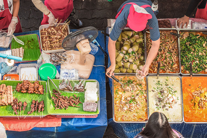 Zu erkennen ist ein gut besuchter Streetfood-Stand mit zahlreichen frisch zubereiteten Gerichten, Spießen und Gemüse. Mehrere Verkäufer portionieren das Essen direkt aus großen Schalen und Behältern. Reise Spartipps Essen
