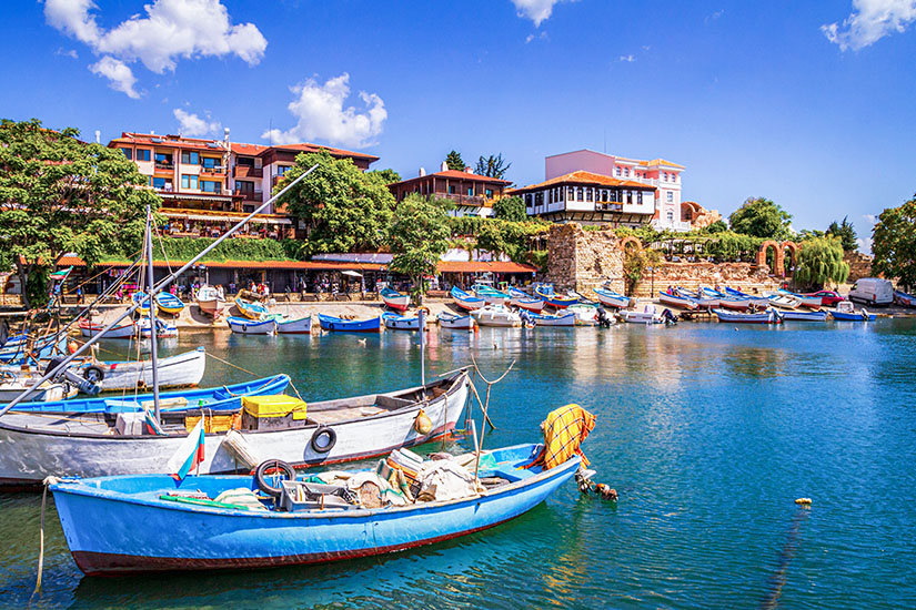 Zu erkennen ist ein malerischer kleiner Hafen mit zahlreichen bunten Fischerbooten und traditionellen Häusern am Ufer. Historische Steinmauern und üppige Vegetation verleihen der Küstenkulisse einen mediterranen Charakter. Urlaub Spartipps Bulgarien