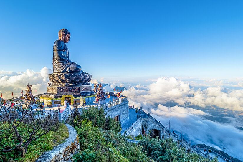 Zu erkennen ist eine riesige sitzende Buddha-Statue auf einer hoch gelegenen Tempelanlage. Wolken ziehen unterhalb des Berggipfels vorbei und verleihen der Szenerie eine eindrucksvolle, fast schwebende Atmosphäre. Billig Reisen Tipps Vietnam