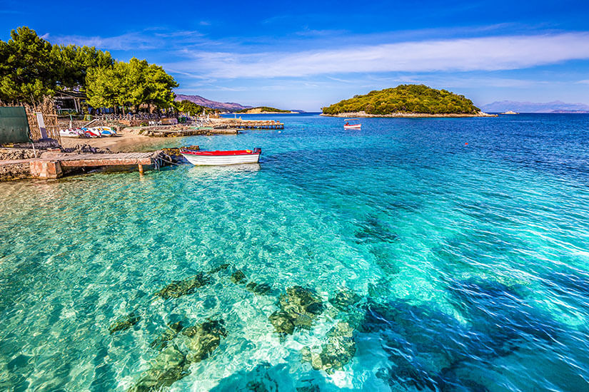 Zu erkennen ist eine idyllische Küstenlandschaft mit kristallklarem, türkisfarbenem Meer und kleinen Booten. Im Hintergrund liegt eine bewaldete Insel, während sich am Ufer ein kleiner Strand mit Bäumen erstreckt. Low Budget Reisen Albanien