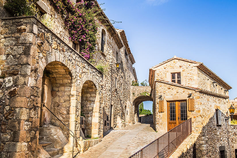 Ein gepflasterter Weg führt durch die Altstadt von Pals vorbei an historischen Steinhäusern zu einem Torbogen. Pals Spanien Altstadt