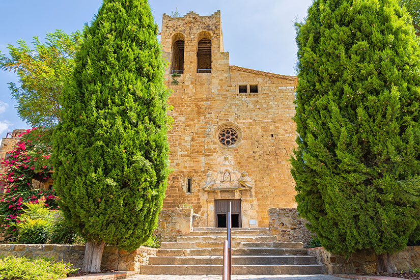 Die Kirche Església de Sant Pere aus hellem Stein ist zwischen zwei grünen Bäumen zu sehen. Pals Spanien Kirche