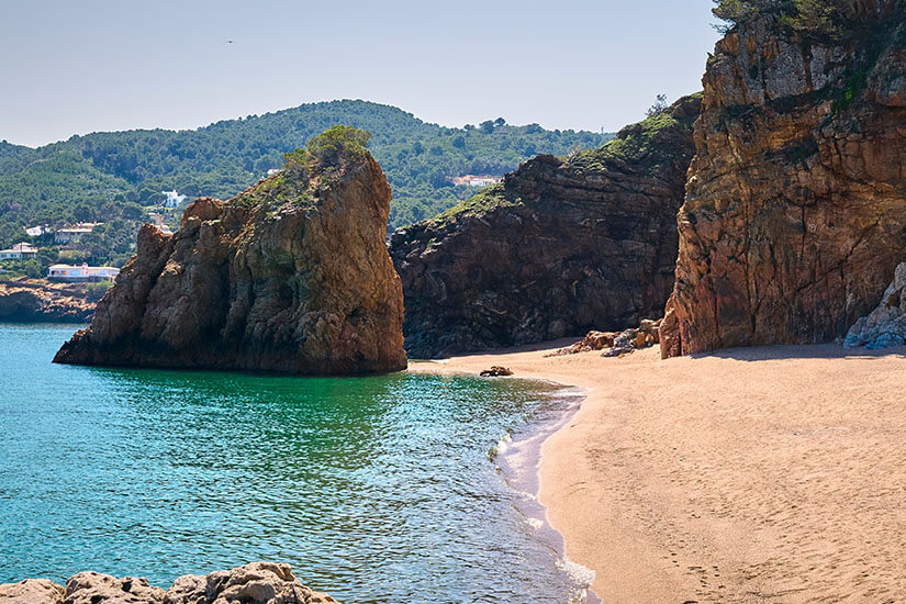 Ein Strandabschnitt mit hellem Sand und klarem Meerwasser ist zu sehen. Große Felsen säumen ihn. Pals Spanien Strand