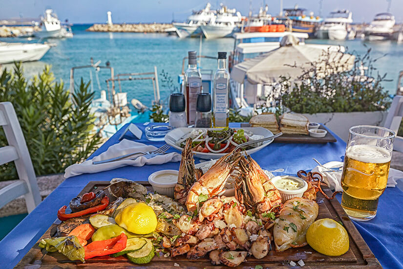 Zu erkennen ist eine üppige Meeresfrüchteplatte mit Garnelen, Tintenfisch, Fisch, gegrilltem Gemüse und Zitronen. Im Hintergrund liegen zahlreiche Boote in einem sonnigen Hafen, daneben steht ein Glas Bier. Zypern Essen Restaurant