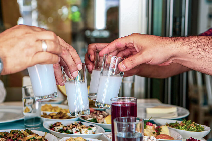 Mehrere Personen stoßen mit Gläsern an, die mit einem milchig-weißen Getränk gefüllt sind. Auf dem Tisch stehen zahlreiche mediterran wirkende Speisen und kleine Vorspeisen. Typisch Zypern Essen Traditionen