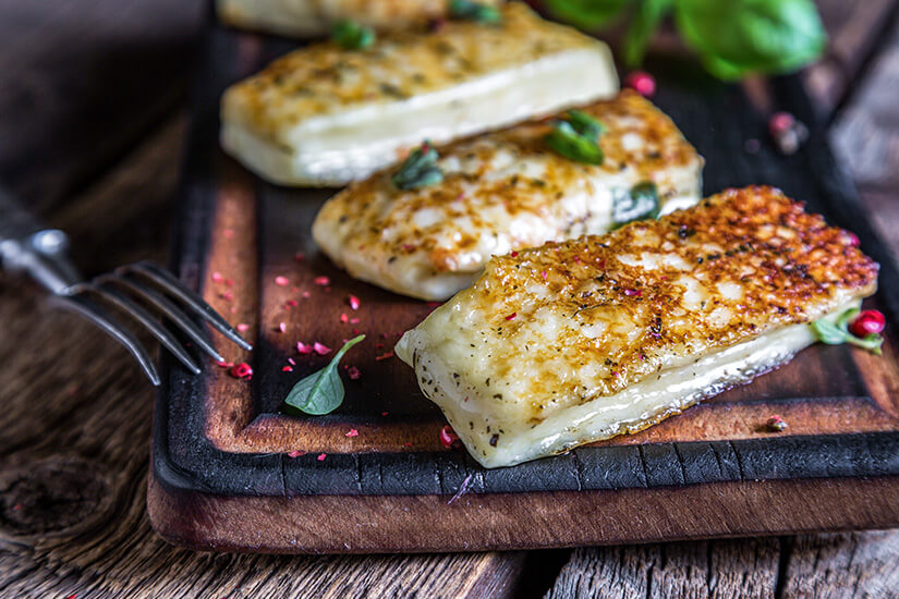 Zu erkennen sind mehrere goldbraun gebratene Scheiben Halloumi auf einem rustikalen Holzbrett. Kräuter und Gewürze ergänzen die appetitlich angerichteten Käsestücke. Zypern Spezialitaeten Halloumi