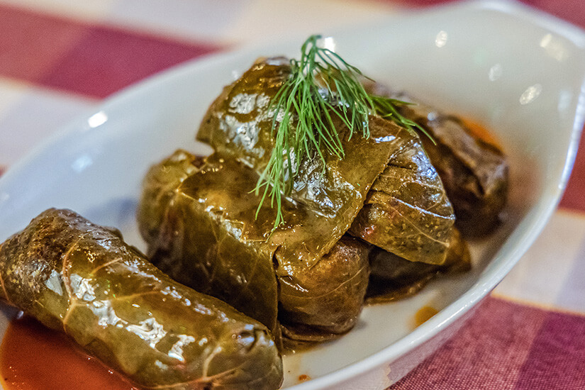 Zu erkennen sind mehrere gefüllte Weinblätter, die in einer kleinen Schale angerichtet sind. Ein Zweig frischer Dill dient als Garnitur. Zypern Kueche Koupepia