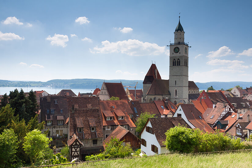 Das Münster St. Nikolaus ragt über die roten Dächer der Altstadt von Überlingen auf. Im Hintergrund sind Hügel und der Bodensee zu sehen. Ueberlingen Deutschland Altstadt