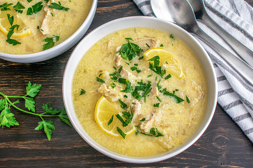 Zu erkennen ist eine cremige Zitronensuppe mit Fleischstücken, garniert mit frischer Petersilie und Zitronenscheiben. Daneben steht eine zweite Schale derselben Suppe. Zypern Essen Avgolemono