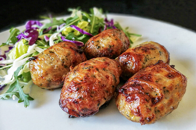 Zu erkennen sind mehrere gegrillte, kräftig gebräunte Fleischröllchen. Dazu wird ein frischer Salat mit verschiedenen Blattsalaten und Rotkohl serviert. Zypriotisches Essen Sheftalia