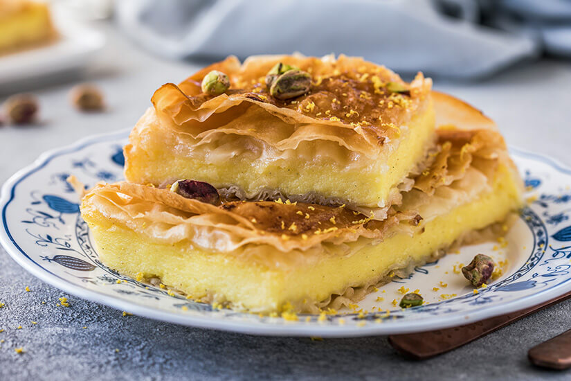 Zu erkennen sind zwei Stücke eines goldbraun gebackenen Desserts aus knusprigem Filoteig und einer cremigen Füllung. Pistazien und feine Zitrusschalen verzieren das Gebäck. Zypern Kueche Galaktoboureko