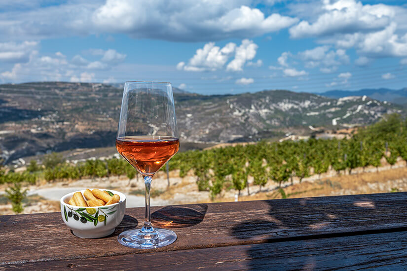 Zu erkennen ist ein Glas Roséwein auf einem Holztisch mit einer kleinen Schale Knabbergebäck daneben. Im Hintergrund erstrecken sich Weinberge über eine hügelige Landschaft unter blauem Himmel. Zypern Essen Wein