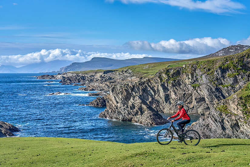 Auf einer sattgrünen Wiese hoch über der Küste radelt eine Person in roter Jacke mit Helm entlang der Klippenkante. Schoenste Radwege der Welt Irland