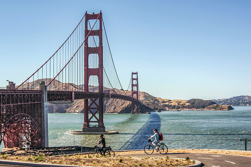 Zwei Radfahrer rollen über einen asphaltierten Uferweg, während sich hinter ihnen die rostrote Golden Gate Bridge mit ihren markanten Pfeilern und Tragseilen über die Bucht spannt. Schoenste Radwege der Welt Kalifornien
