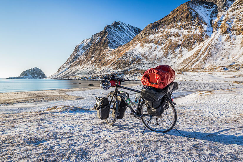 Schwer bepackt mit Satteltaschen und einem knallroten Packsack steht ein Reiserad allein auf einem schneebedeckten Strand am Fjord. Schoenste Radwege der Welt Norwegen
