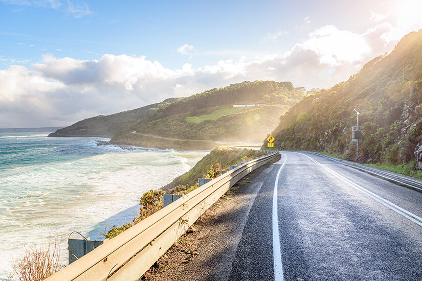 Direkt an der Steilküste windet sich eine asphaltierte Straße mit Leitplanke um bewaldete grüne Hügel, im Gegenlicht der tief stehenden Sonne. Schoenste Radwege der Welt Australien