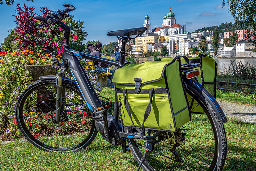 Mit knallgrünen Doppelpacktaschen am Gepäckträger steht ein E-Bike auf einer Wiese neben einem bunten Blumenbeet direkt am Fluss. Schoenste Radwege der Welt Oesterreich