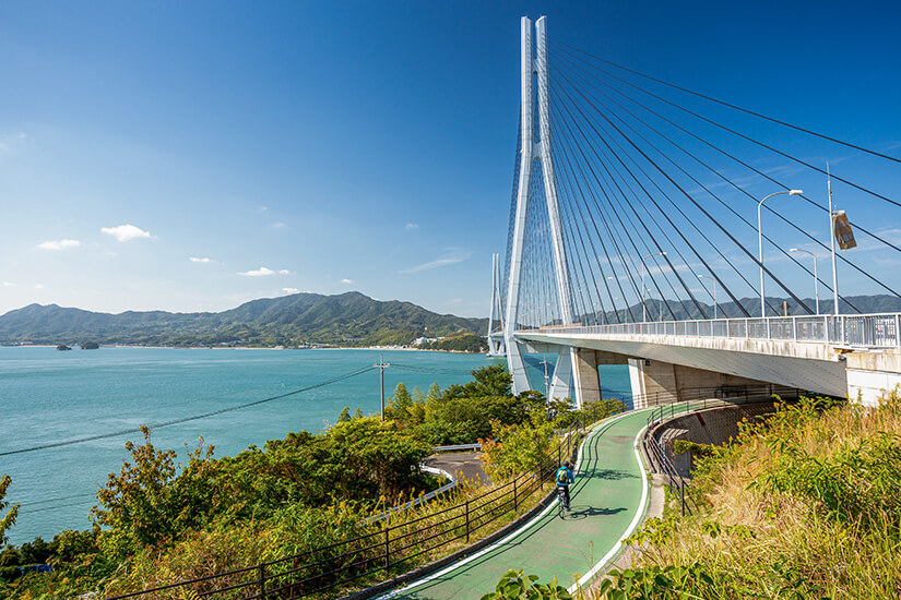 Auf einer grün markierten Radspur schlängelt sich ein einzelner Fahrer die Rampe zu einer weißen Schrägseilbrücke hinauf, die sich weit über die türkisblaue Meerenge spannt. Schoenste Radwege der Welt Japan