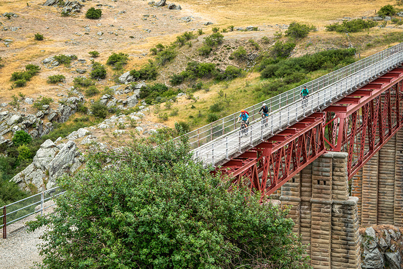 Drei Radfahrer überqueren hintereinander eine rote Stahlfachwerkbrücke auf massiven Steinpfeilern, die über eine Schlucht führt. Schoenste Radwege der Welt Neuseeland