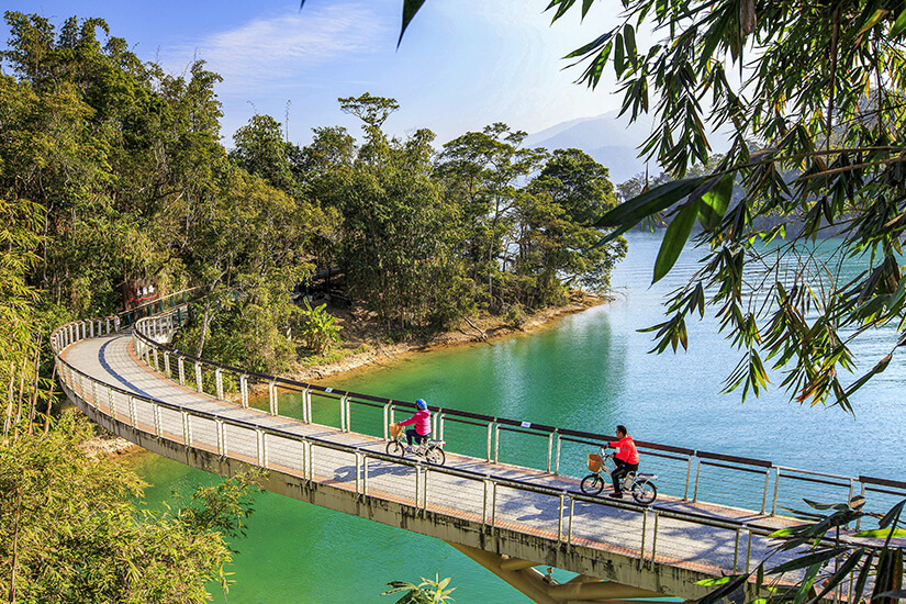 Zwei Radfahrer in roten Jacken rollen über einen geschwungenen Radweg, der auf Stelzen dicht über dem türkisgrünen Wasser eines Sees entlangführt. Schoenste Radwege der Welt Taiwan