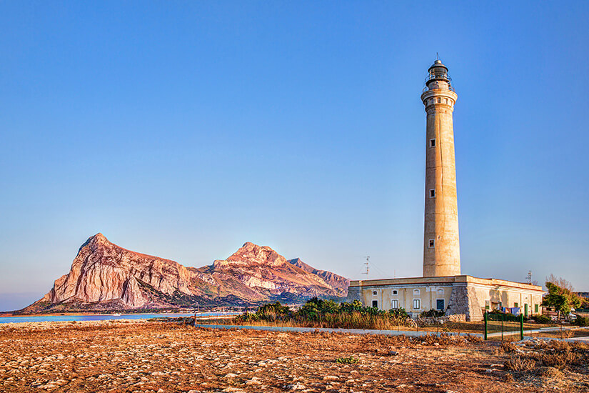 Ein hoher, schlanker Leuchtturm ragt auf einer steinigen Küstenebene in San Vito Lo Capo empor. An seinem Fuß steht ein flaches, beige-farbenes Gebäude. Im Hintergrund erheben sich im warmen Sonnenlicht markante, schroffe Bergfelsen vor einem klar emporragenden, blauen Himmel. San Vito Lo Capo Italien Faro