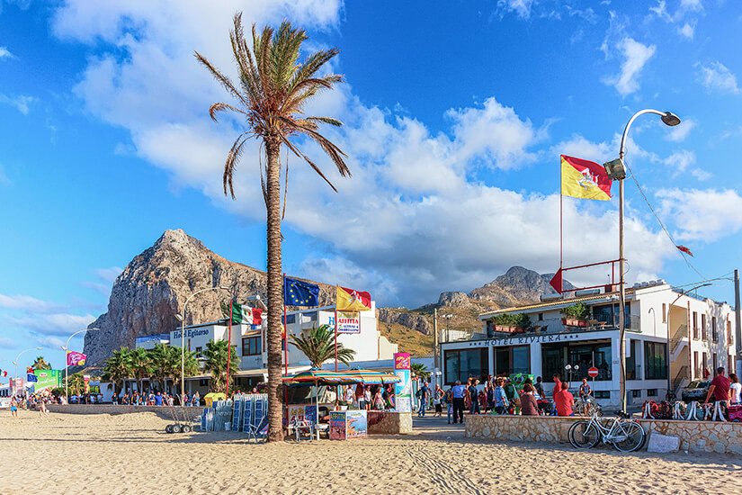 Blick auf die sonnige Promenade am Sandstrand von San Vito Lo Capo. Eine hohe Palme ragt vor weiß getünchten Hotelgebäuden und kleinen Verkaufsständen in den blauen Himmel mit leichten Wolken. Im Hintergrund erhebt sich der imposante, felsige Monte Monaco. San Vito Lo Capo Italien Spiaggia