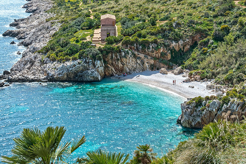 Spektakuläre Luftaufnahme der Bucht Cala Tonnarella dell’Uzzo im Naturschutzgebiet Zingaro nahe San Vito Lo Capo. Kristallklares, türkisblaues Wasser trifft auf einen hellen Kiesstrand, der von steilen, grün bewachsenen Felsen umrahmt wird. Auf den Klippen oberhalb der Bucht steht ein kleines, historisches Steingebäude. San Vito Lo Capo Italien Cala Tonnarella
