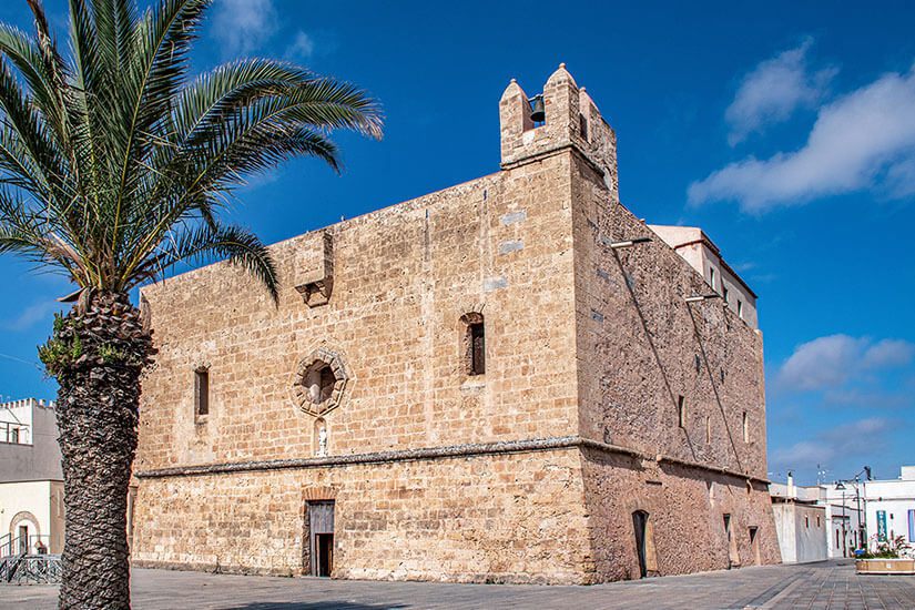 Die festungsartige Kirche Santuario di San Vito Martire aus hellen Natursteinen in San Vito Lo Capo. Das wuchtige Bauwerk besitzt einen kleinen Glockenturm an der Ecke, eine Rosette über dem Portal und wenige schmale Fenster. Links im Vordergrund steht eine große Palme vor dem sonnigen, blauen Himmel. San Vito Lo Capo Italien Santuario di San Vito Martire