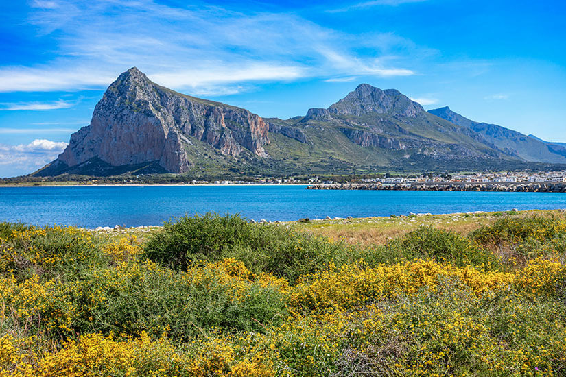 Blick über eine hügelige Wiese voller gelber Ginsterblüten auf das tiefblaue Meer bei San Vito Lo Capo. Am gegenüberliegenden Ufer erheben sich die markanten, schroffen Felsmassive des Monte Monaco unter einem leicht bewölkten, strahlend blauen Himmel. San Vito Lo Capo Italien Monaco