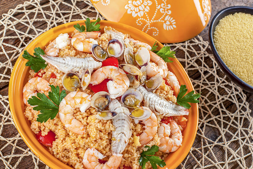 Ein gelber Servierteller gefüllt mit traditionellem sizilianischem Couscous mit Meeresfrüchten. Auf dem körnigen Couscous liegen rosa Garnelen, Muscheln in der Schale, weiße Fischfilets sowie frische Petersilie und Tomatenstücke. Der Teller steht auf einer geflochtenen Unterlage neben einer kleinen Schale mit ungekochtem Couscous und einem passenden gelben Keramikdeckel. San Vito Lo Capo Italien Cous Cous