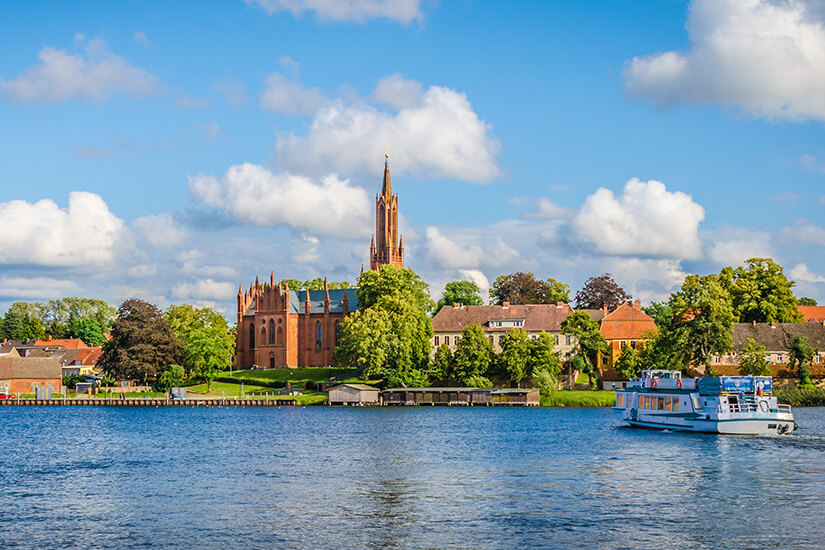 Zu erkennen ist eine idyllische Uferlandschaft mit einer markanten neugotischen Backsteinkirche, umgeben von Bäumen und Häusern. Auf dem See fährt ein weiß-blaues Ausflugsschiff unter einem sonnigen, leicht bewölkten Himmel. Seenplatte Mecklenburg Region