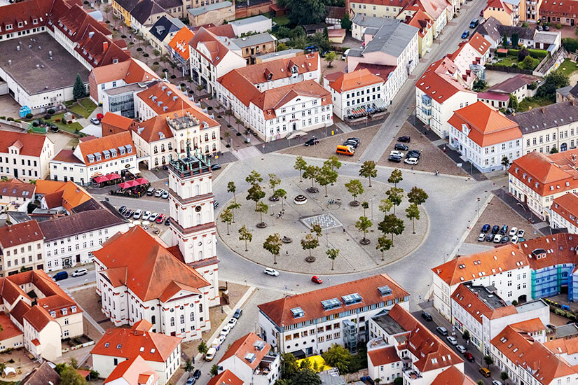 Zu erkennen ist eine Luftaufnahme des zentralen Marktplatzes von Neustrelitz mit seiner charakteristischen kreisförmigen Anlage. Rund um den Platz stehen historische Gebäude mit roten Dächern sowie eine markante Kirche mit hohem Turm. Mecklenburgische Seenplatte Neustrelitz