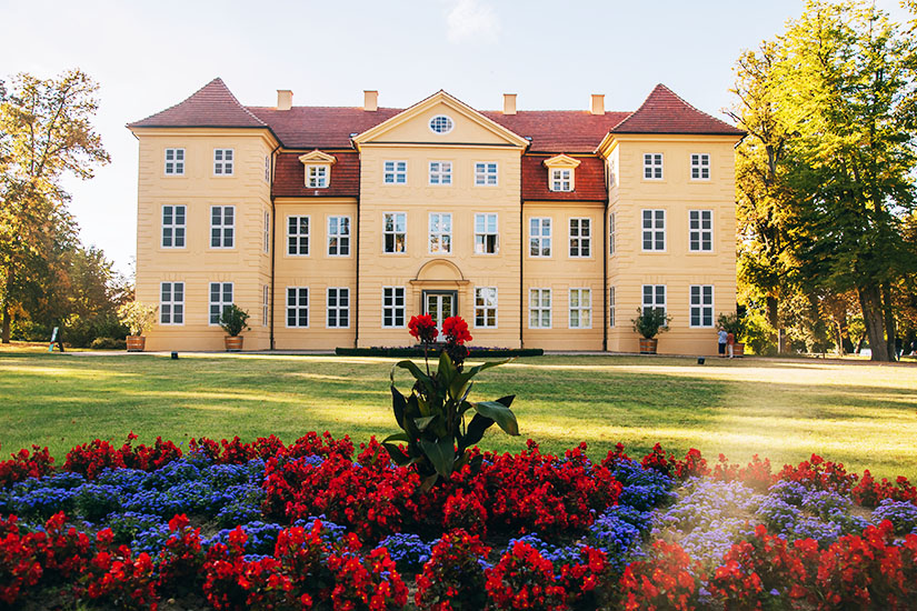 Zu erkennen ist Schloss Mirow mit seiner hellgelben Fassade und dem roten Ziegeldach, umgeben von einer gepflegten Parkanlage. Im Vordergrund sorgt ein farbenprächtiges Blumenbeet mit roten und blauen Blüten für einen dekorativen Akzent. Mecklenburg-Vorpommern Schloss Mirow