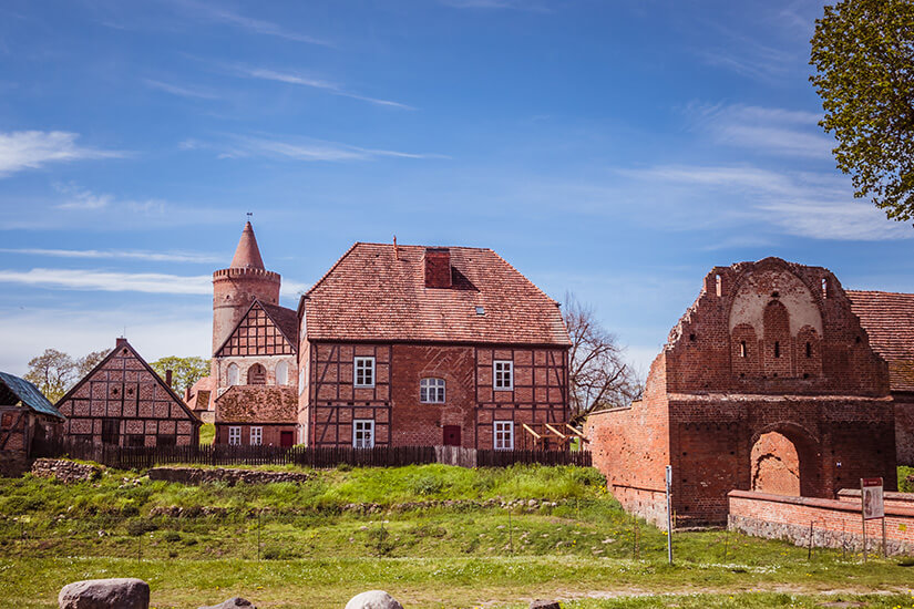 Zu erkennen ist eine historische Burganlage mit Fachwerkhäusern, einem runden Turm und alten Backsteinmauern. Teilweise erhaltene Ruinen und die traditionelle Architektur verleihen der Anlage einen mittelalterlichen Charakter. Mecklenburger Seenplatte Burg