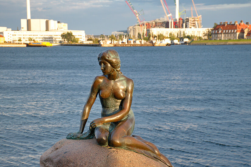 Die Statue "die kleine Meerjungfrau" thront auf einem Felsen im Wasser. Im Hintergrund sind Gebäude und Hafenteile von Kopenhagen zu sehen. Kopenhagen Sehenswuerdigkeiten Meerjungfrau
