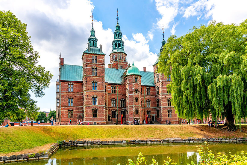 Das Schloss Rosenborg mit seiner Roten Fassade und den grünen Dächern ragt vor einem Gewässer auf. Kopenhagen Sehenswuerdigkeiten Rosenborg