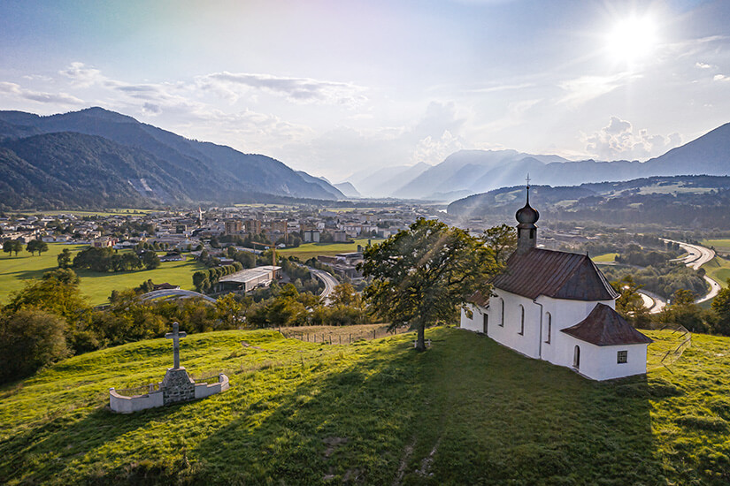 Eine kleine weiße Kapelle steht auf einem sattgrünen Hügel hoch über dem Tiroler Unterinntal. Der Blick schweift über die Dächer von Wörgl bis hin zu den umliegenden Bergketten, während die Abendsonne die Landschaft in goldenes Licht taucht. Wörgl Österreich Kapelle