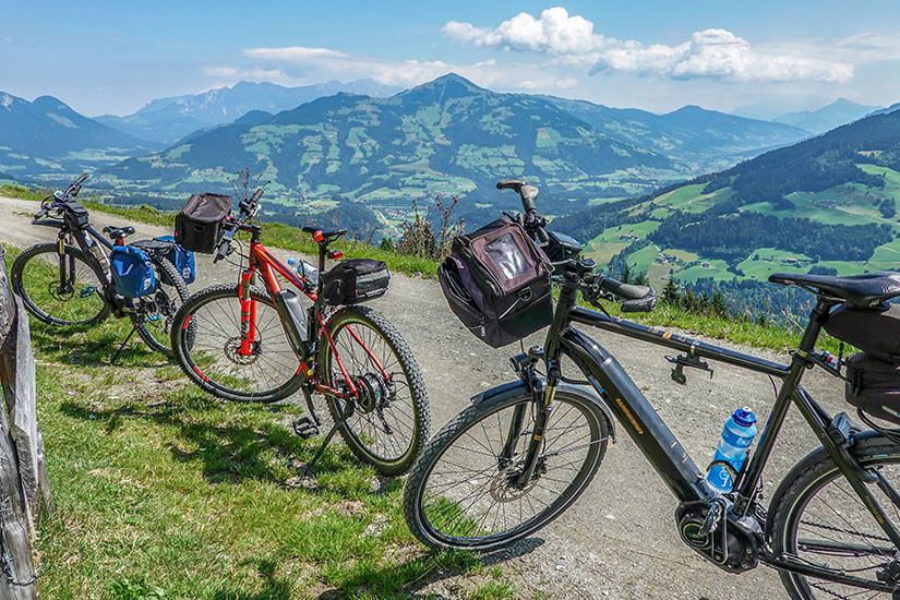 Drei bepackte Mountainbikes stehen am Wegesrand einer Bergstraße mit freiem Blick auf die sanften Hügel und Gipfel der Kitzbüheler Alpen. Die saftig grünen Almwiesen und das strahlende Sommerwetter laden zu ausgedehnten Touren durch die Region ein. Wörgl Österreich Radtour