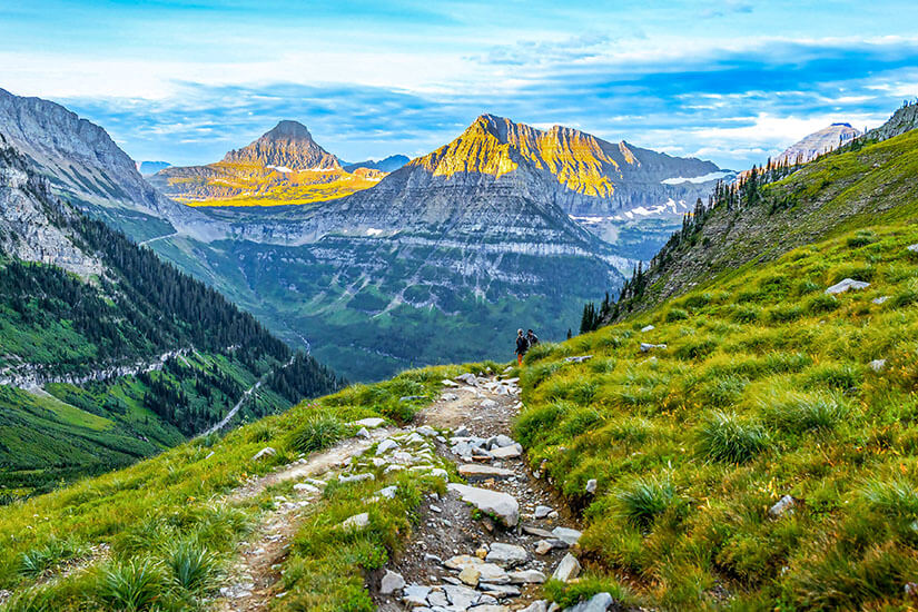 Ein steiniger Wanderpfad schlängelt sich durch saftig grüne Bergwiesen einen Hang entlang, auf dem zwei winzige Wanderer unterwegs sind. Bedrohte Reiseziele Klimawandel USA