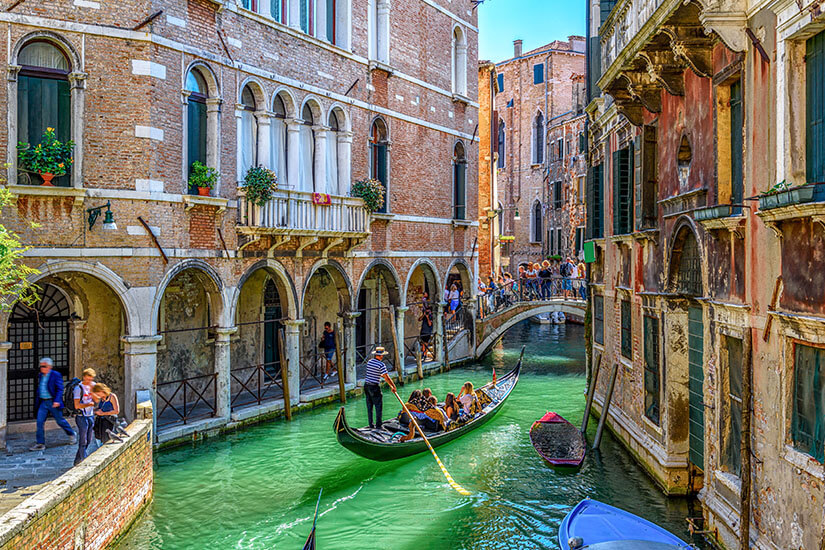 Auf grün schimmerndem Wasser stakt ein Gondoliere mit Strohhut seine besetzte Gondel durch einen schmalen venezianischen Kanal, gesäumt von verwitterten Backsteinfassaden mit Arkaden, Blumenbalkonen und Bogenfenstern. Bedrohte Reiseziele Klimawandel Venedig