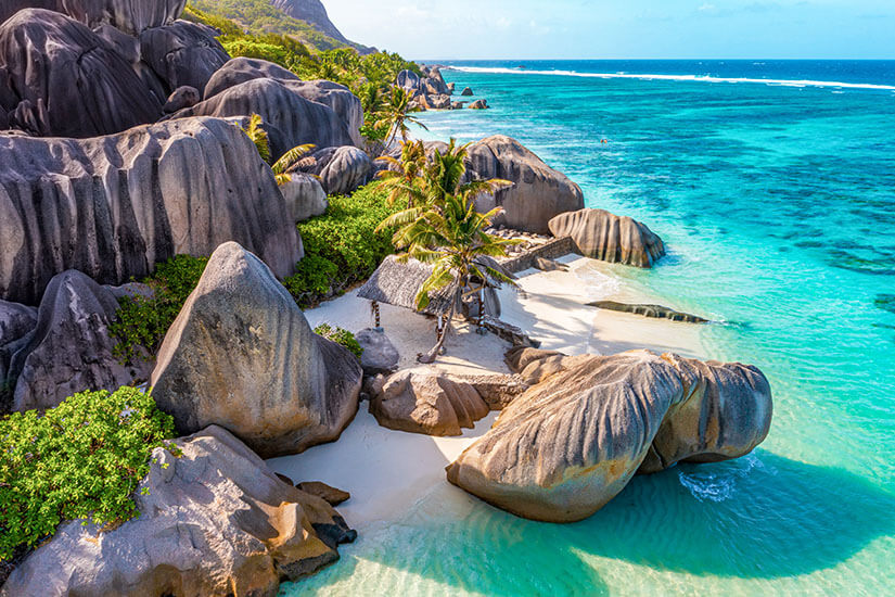 Von oben blickt man auf mächtige, glatt geschliffene Granitfelsen, die eine winzige weiße Sandbucht mit Palmen und einer strohgedeckten Hütte einrahmen – die berühmte Anse Source d'Argent auf La Digue. Bedrohte Reiseziele Klimawandel Seychellen