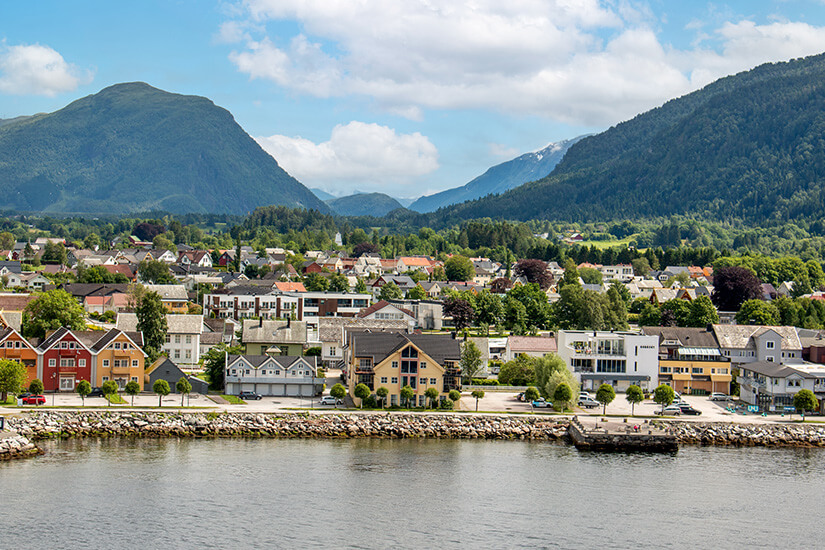 Zu erkennen ist die norwegische Ortschaft Nordfjordeid direkt am Wasser, umgeben von hohen, dicht bewaldeten Bergen. Entlang des Ufers stehen zahlreiche Häuser, während sich die Siedlung weit ins grüne Tal hineinzieht. Nordfjordeid Stadt