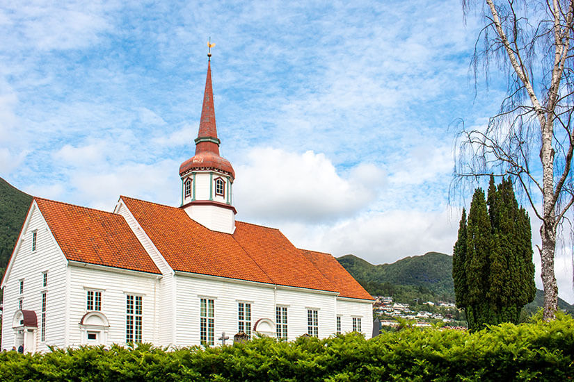 Zu erkennen ist eine weiße Holzkirche mit rotem Ziegeldach und einem hohen, schlanken Kirchturm. Im Hintergrund erheben sich grüne Berge, während Bäume und Sträucher das Gebäude umgeben. Nordfjordeid Kirche