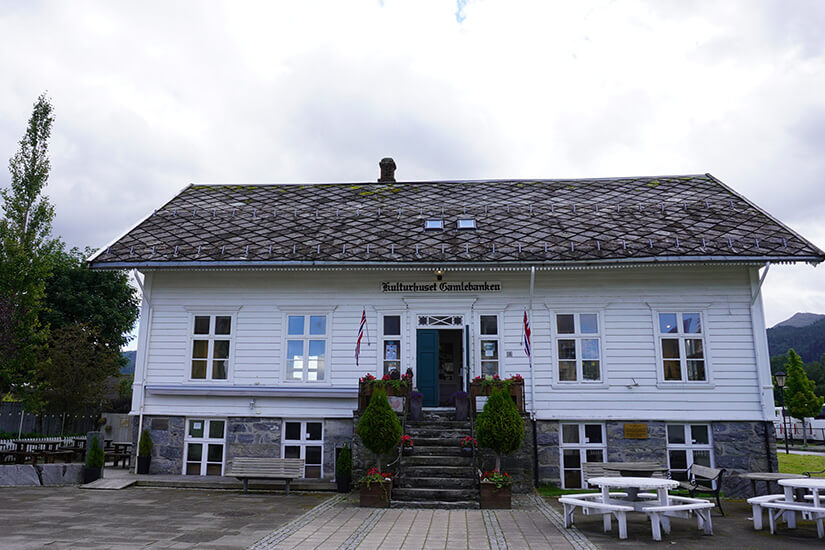 Zu erkennen ist ein traditionelles weißes Holzgebäude mit Steinsockel und norwegischen Flaggen am Eingang. Über der Tür steht „Kulturhuset Gamlebanken“, davor befinden sich Sitzbänke und Picknicktische. Nordfjordeid Kulturhaus